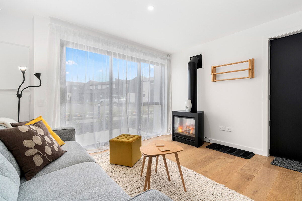 A comfortable living space is presented, featuring a light grey sofa adorned with decorative pillows. A small wooden table is positioned in front, accompanied by a yellow ottoman. A contemporary fireplace is positioned against the wall, with soft natural light filtering through sheer curtains.