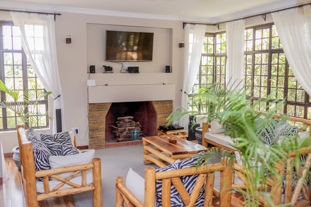 A cozy living area is furnished with natural wooden seating featuring striped cushions. A stone fireplace stands at one end, complemented by a wall-mounted television. Large windows are draped with sheer curtains, allowing soft light to illuminate the space and potted plants to add a touch of greenery.