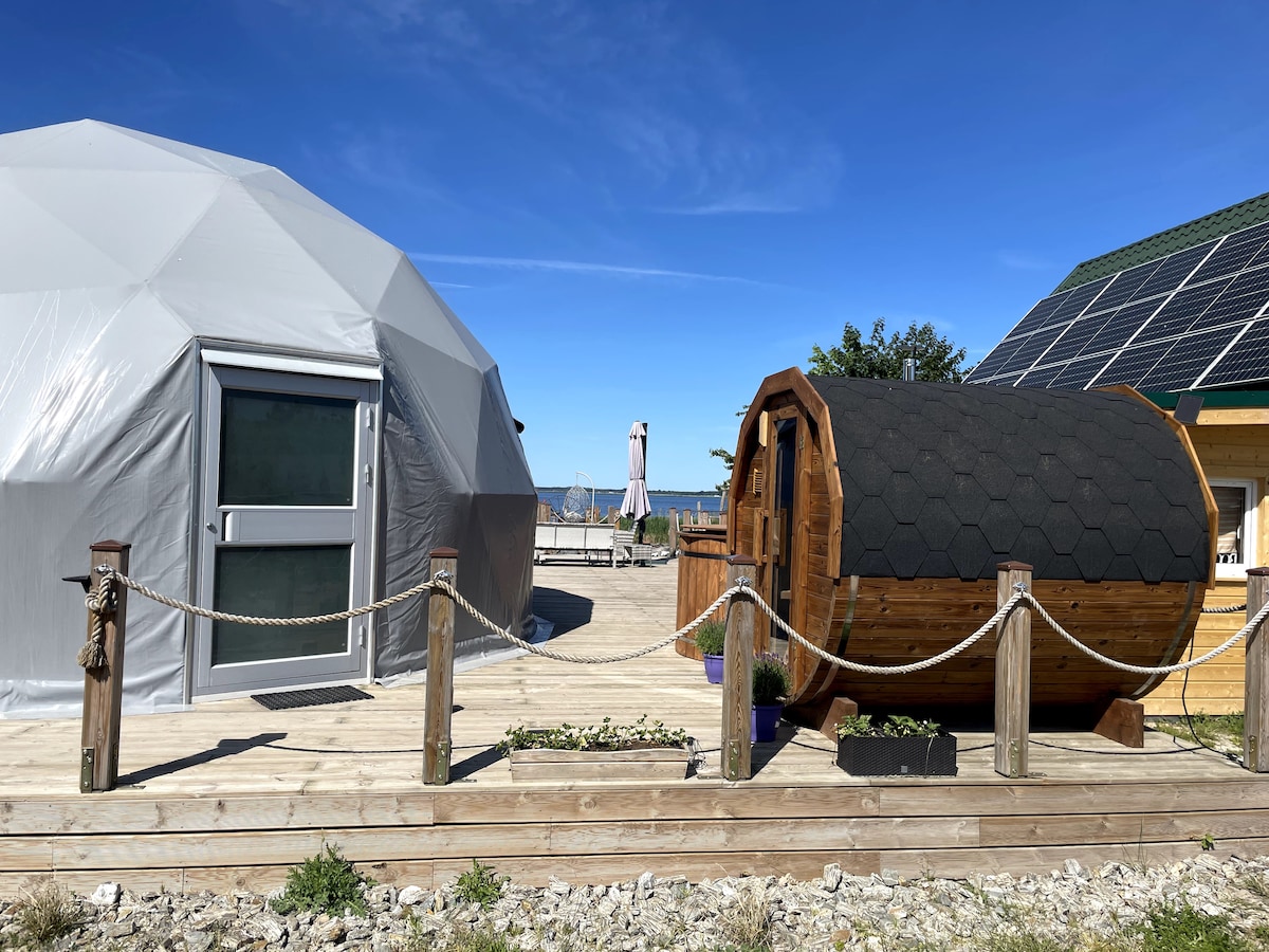 A spacious, geodesic dome structure is positioned alongside a wooden sauna. Sunlight brightens the scene, highlighting the surrounding wooden deck and tranquil outdoor area, with glimpses of the lake visible in the background.
