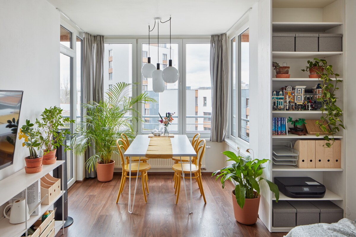 A brightly lit dining area features a white table surrounded by three yellow chairs. Large windows allow natural light to fill the space, with green plants placed strategically around the room. A bookshelf displays various decorative items and storage boxes.