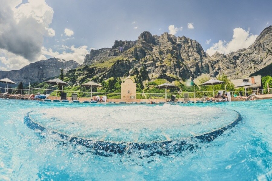 A sparkling pool is framed by majestic mountains under a clear sky. Umbrellas provide shade for lounge chairs along the pool's edge, inviting relaxation. The shimmering water features a soft wave pattern, creating a tranquil atmosphere for guests.