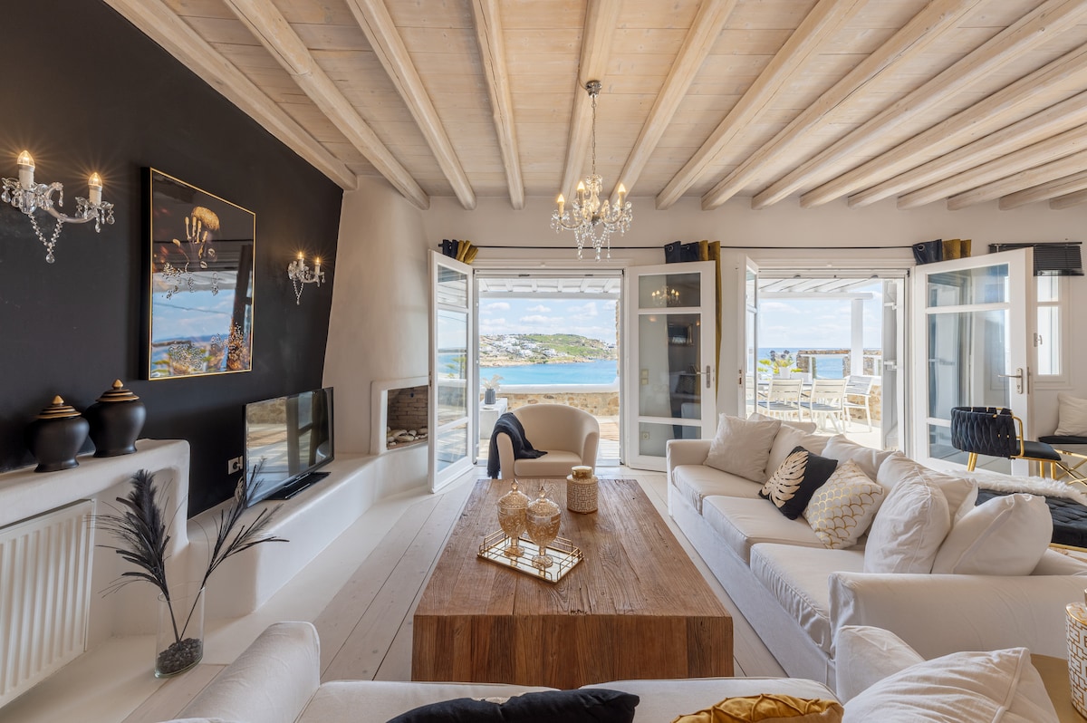 A spacious living area features light-colored sofas arranged around a wooden coffee table. Floor-to-ceiling doors open to reveal a stunning sea view, while the ceiling showcases natural wooden beams and elegant lighting fixtures add a touch of sophistication.