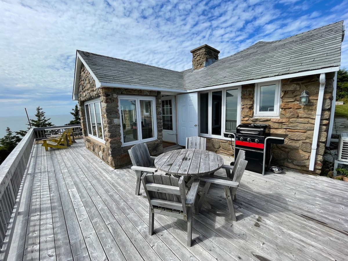 A spacious outdoor deck features a circular wooden table surrounded by several weathered chairs. A gas grill is positioned nearby, and large glass windows offer unobstructed views of the ocean. The stone cottage is seen in the background, complemented by a clear blue sky.