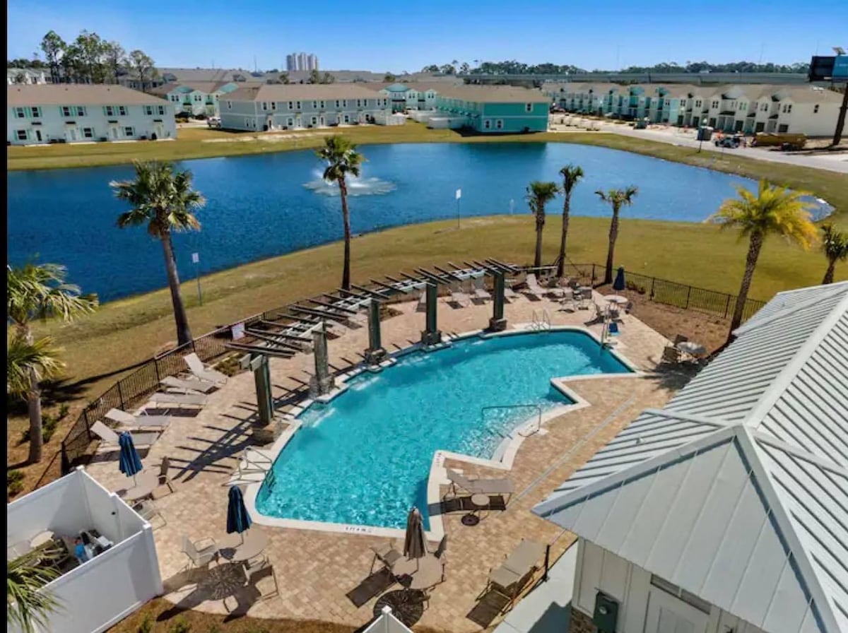 An aerial view of a community pool surrounded by palm trees, featuring lounge chairs and umbrellas. The clear water of the pool sparkles under the sunlight, while a serene lake is visible nearby, enhancing the outdoor atmosphere.