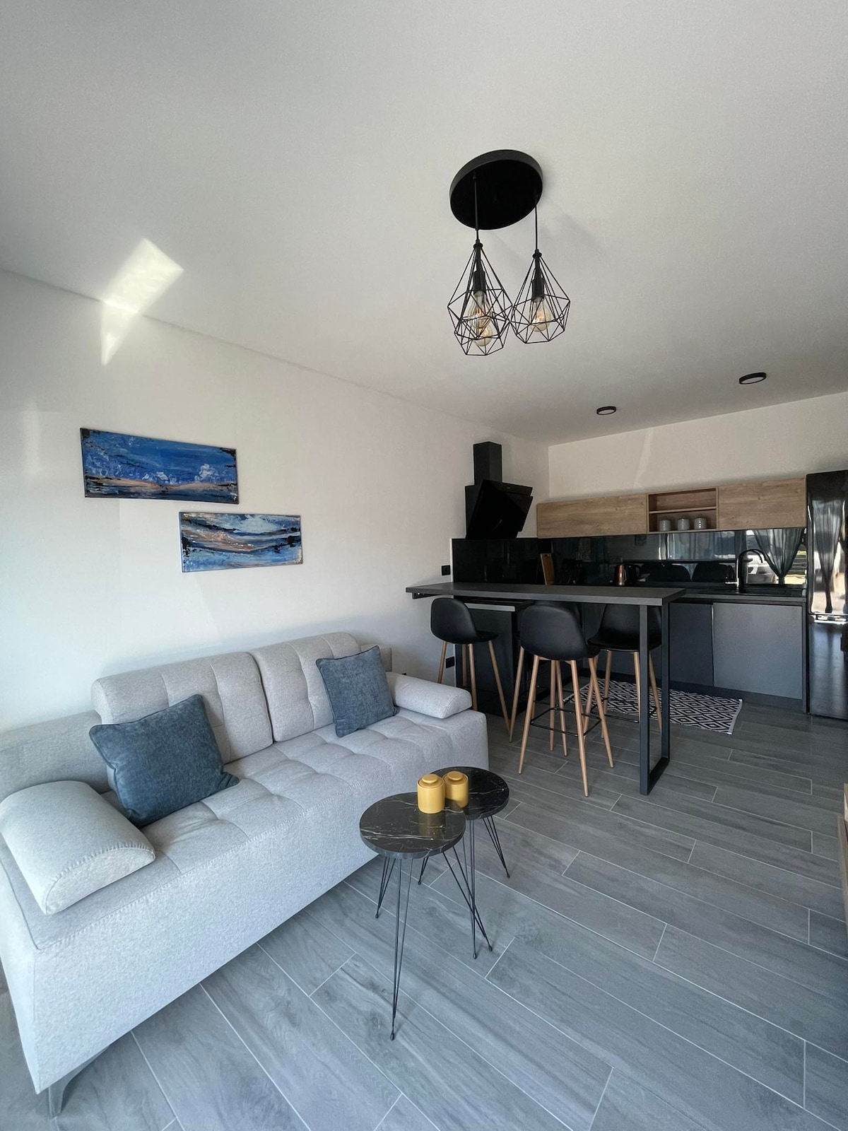 A modern living area features a light-colored sofa with two cushions, positioned near a small black table. Above, a stylish pendant light adds illumination. A sleek kitchen area is visible in the background, complemented by barstool seating and decorative wall art.