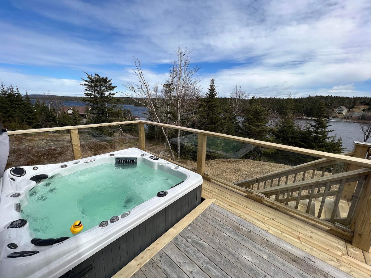 A six-person hot tub is situated on a wooden deck, offering views of the tranquil pond and surrounding trees. The water features a light blue hue, and a small rubber duck floats nearby. The scene is framed by a clear glass railing and a blue sky dotted with clouds.
