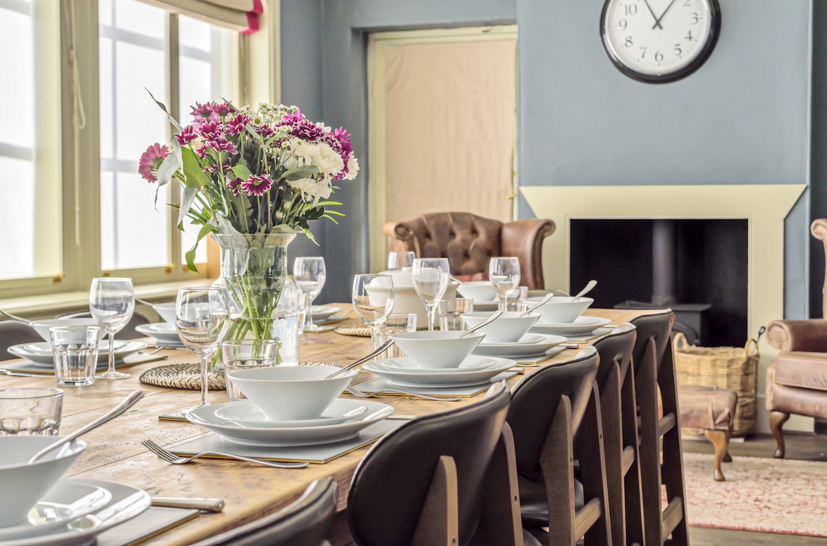 A large dining table is set with elegant dishware, including white plates and glassware. A vase filled with fresh flowers serves as a centerpiece, and soft lighting illuminates the space. A comfortable seating area with a leather armchair is visible in the background.