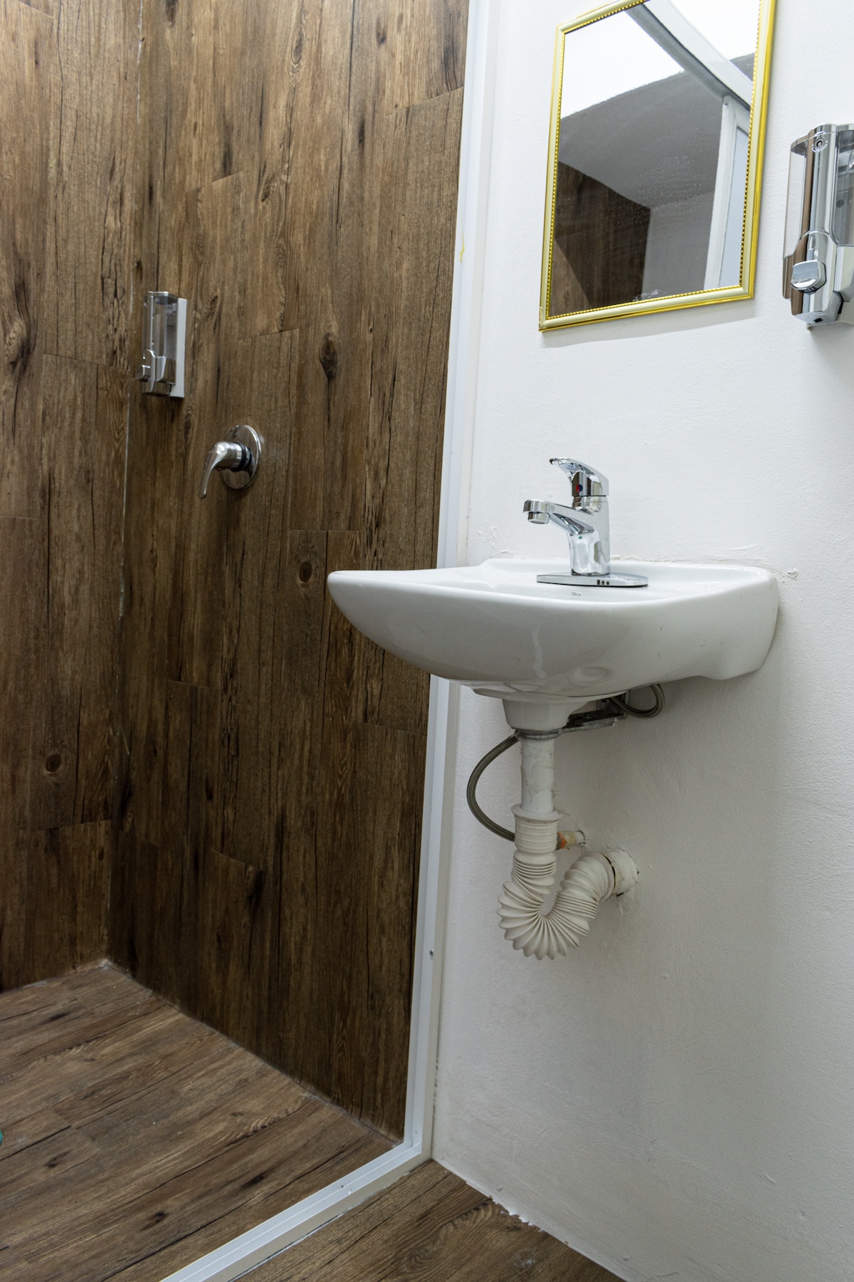A compact bathroom area features a standing shower with a glass enclosure, complemented by a small sink mounted on the wall. The wood-paneled accent wall offers a warm contrast to the white fixtures, while a mirror is positioned above the sink.