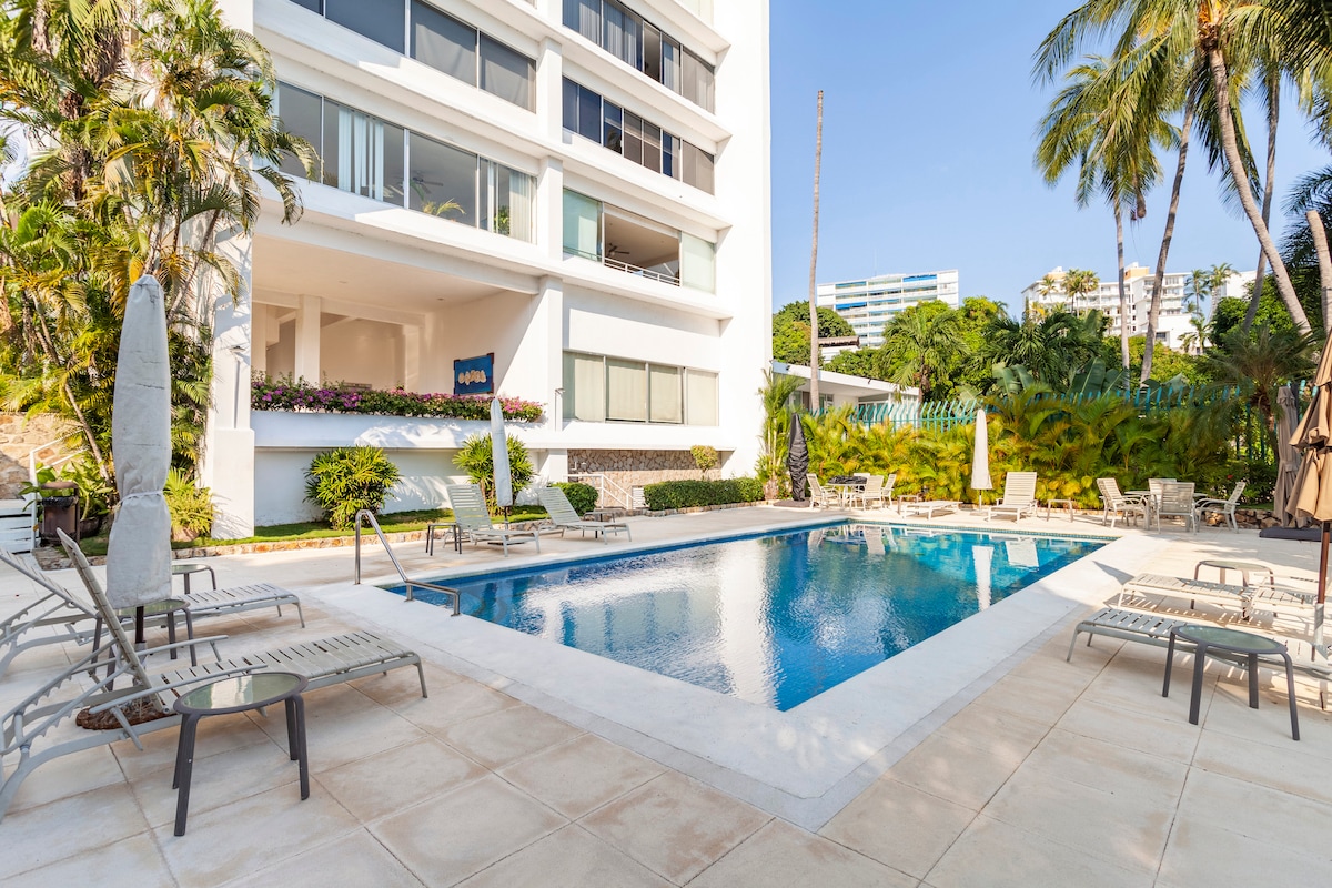 A serene pool area is surrounded by lush greenery and palm trees, featuring comfortable lounge chairs. The clear blue water reflects the sunlight. A modern building with large windows stands in the background, offering an inviting atmosphere for relaxation.