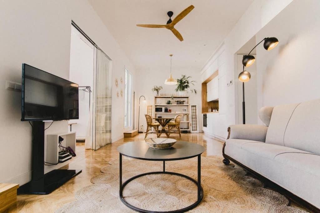 A spacious living area is presented, featuring a minimalist design. A flat screen TV sits on a stand opposite a comfortable sofa. A round coffee table rests on a textured rug, while a dining area with wooden chairs and a large pendant light is visible in the background.