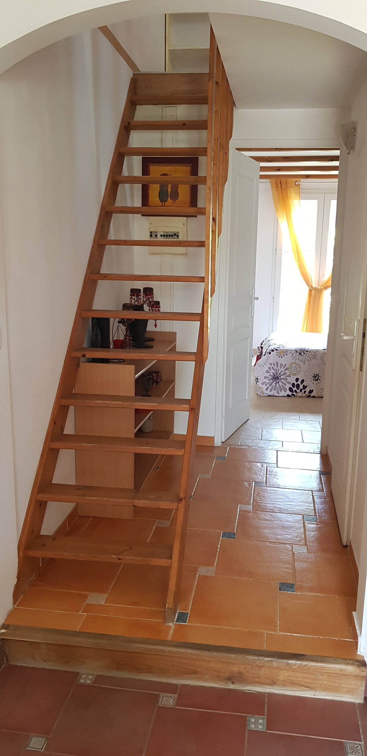 A wooden staircase leads to the upper level, framed by a white archway. The tiled floor offers a warm, earthy tone. A small wooden shelf is positioned near the bottom of the stairs, while a glimpse of a living area can be seen through the open door.