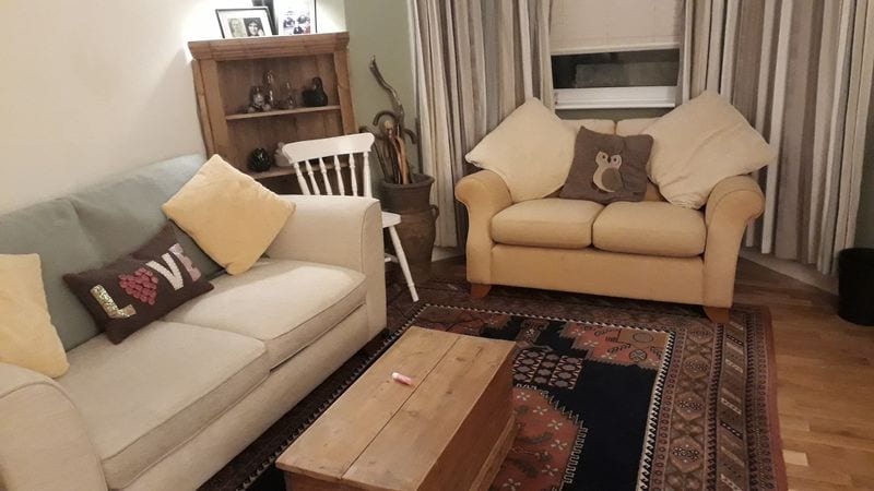 A living room area features a light-colored sofa and a yellow armchair. A wooden coffee table is placed in front, accompanied by a patterned rug. A small bookshelf holds decorative items, and curtains frame the window, allowing for soft natural light.