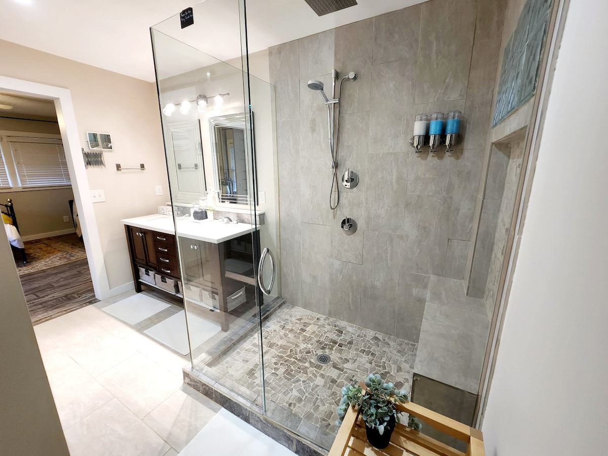 A modern bathroom features a spacious glass-enclosed shower with a rain shower head and stone flooring. A double vanity with under-sink storage is present, along with bright mirrors and light fixtures. A small bench and potted plant add a touch of comfort.