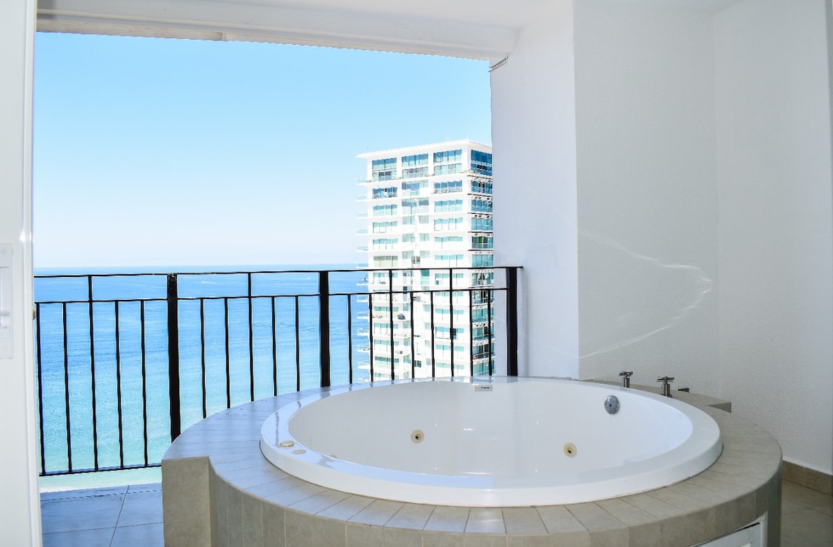 A white, circular jacuzzi is positioned near a large balcony window, offering an unobstructed view of the ocean and sky. The serene waters below reflect the blue hues, while the modern building structure is visible in the background, enhancing the space's bright and airy feel.