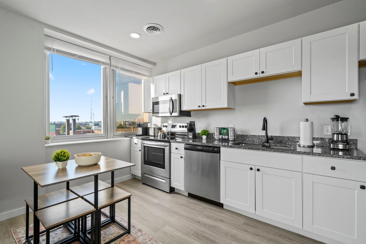A modern kitchen features white cabinetry and stainless steel appliances. A small dining table is visible, accompanied by black metal chairs. Natural light from a window brightens the space, showcasing the granite countertops and an organized array of kitchen utensils.