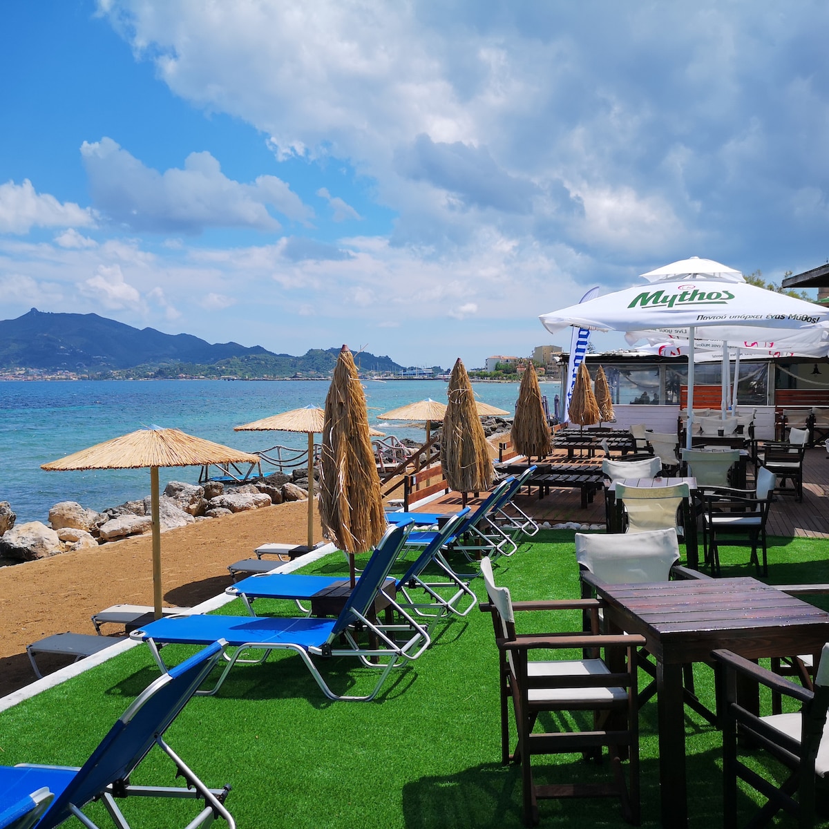 A sunlit beach area features a row of lounge chairs positioned on green grass, shaded by straw umbrellas. Wooden tables and chairs are arranged for guests, with a view of the water and distant hills under a partly cloudy sky.