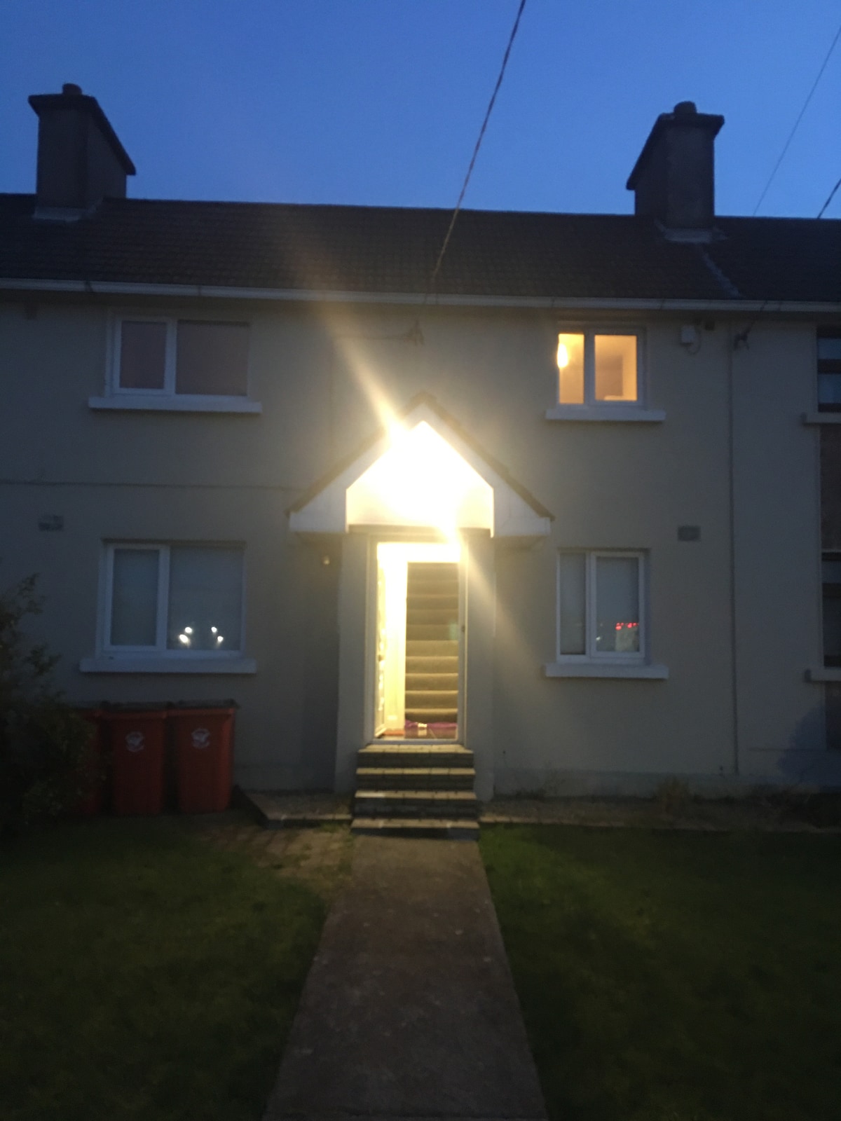 The exterior view of a two-story building is taken during twilight, showcasing a well-lit entrance with steps leading up to a door. Lush grass surrounds the walkway, and two red bins are positioned to the left of the entrance.
