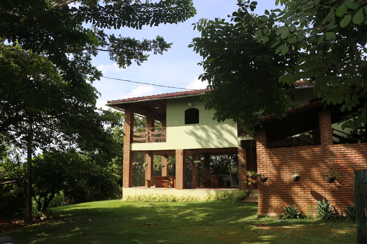 A spacious house is surrounded by lush greenery, featuring a welcoming brick exterior. Large windows allow natural light to fill the interiors. A balcony extends from the upper level, providing outdoor seating and a tranquil view of the landscape.