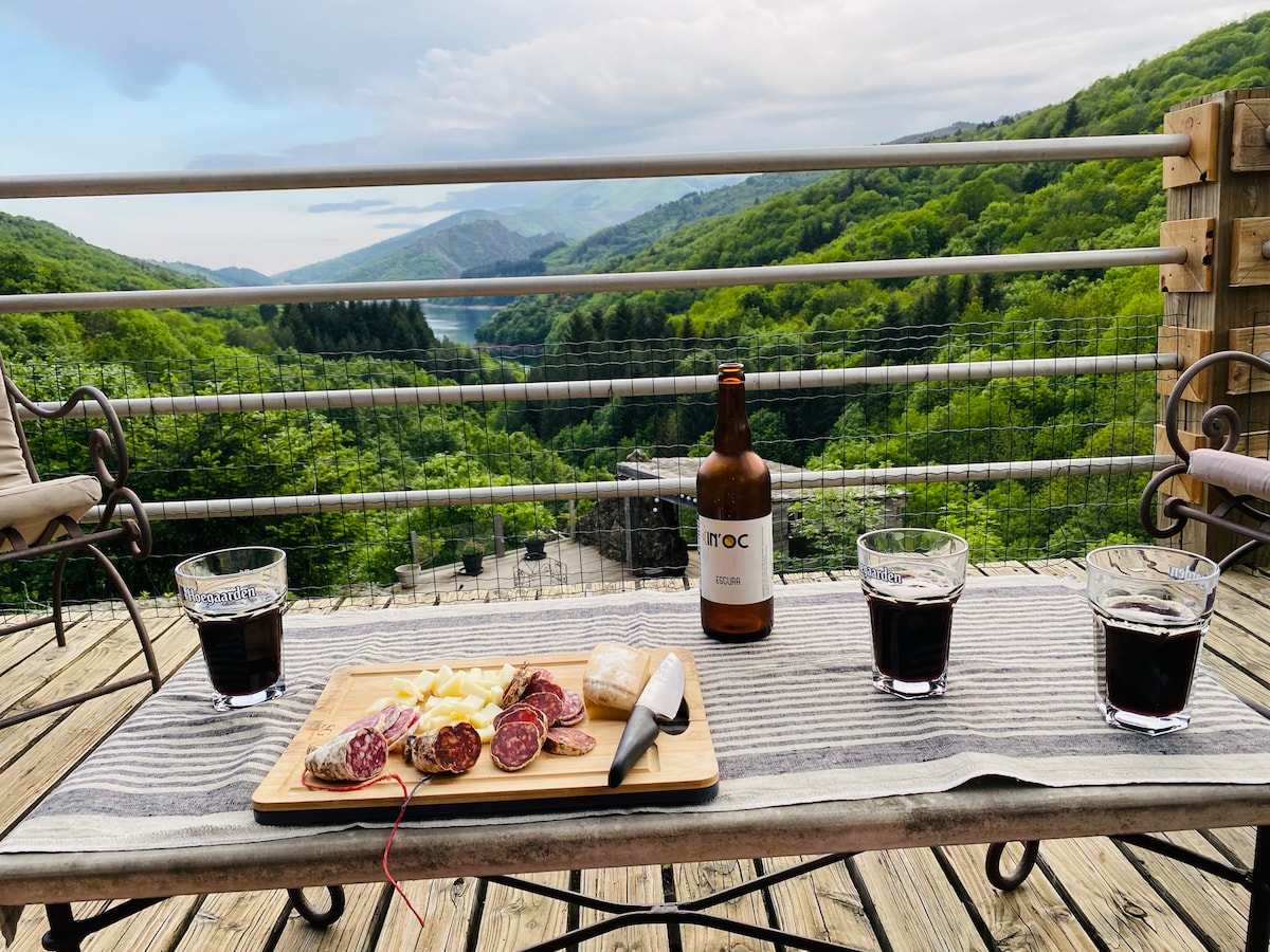 A wooden terrace features a charcuterie board with various cured meats and cheeses, alongside a bottle of beer. Three glasses containing dark beverages are positioned on a striped tablecloth. A scenic view of lush green hills and a lake is visible in the background.