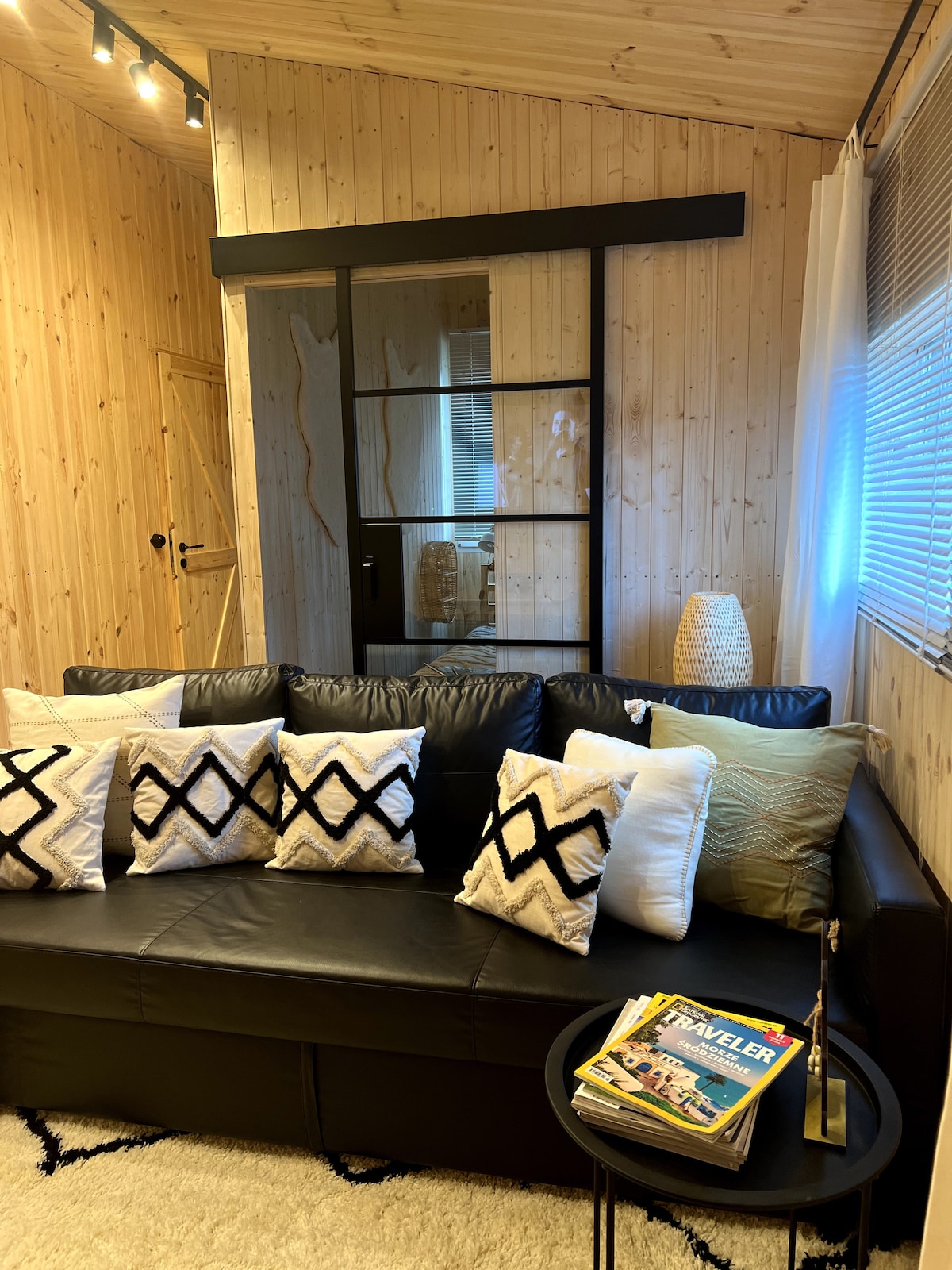 A black sofa adorned with decorative cushions in contrasting patterns is positioned in the room. A small circular table holds magazines, while a woven light fixture and warm wood paneling create a cozy ambiance.
