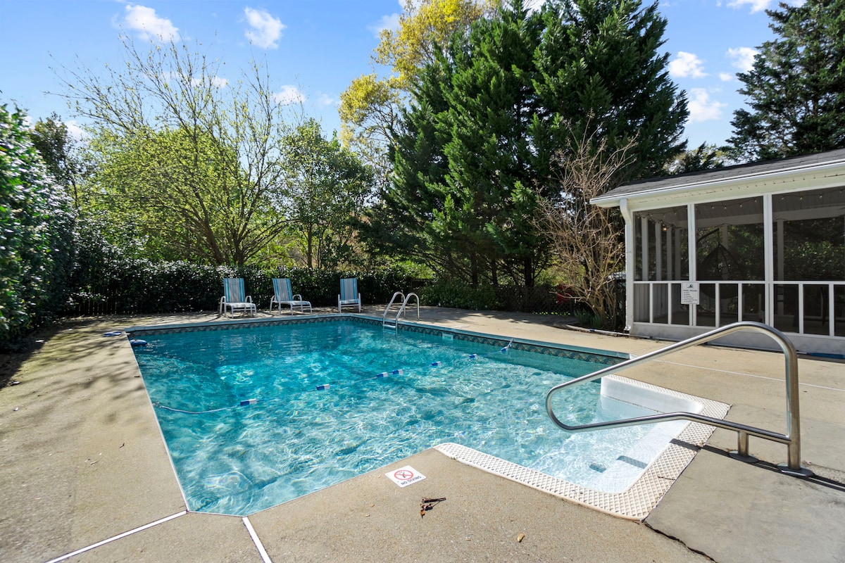 The inviting pool area features a clear, sparkling swimming pool surrounded by a spacious concrete patio. Lounge chairs are positioned nearby, and hedges provide privacy. Sunlight filters through the trees, creating a calming outdoor environment.