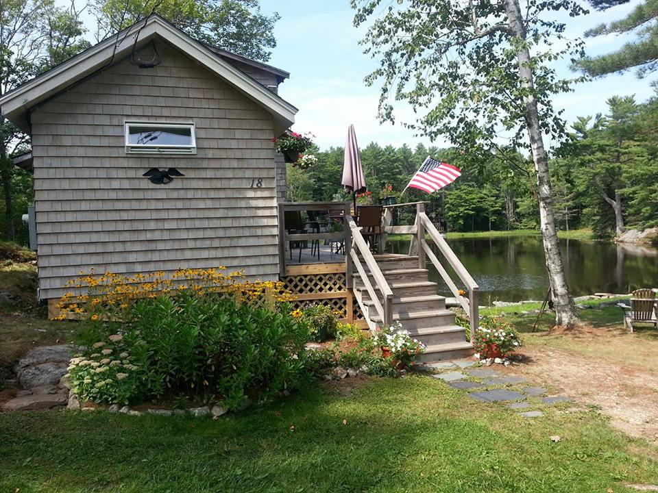 The exterior of Gull Cottage is shown, featuring natural wood siding and a welcoming front porch. Stairs lead up from the garden area adorned with colorful flowers towards the deck, which has an umbrella providing shade. Beals Pond is visible behind the cottage, surrounded by trees.