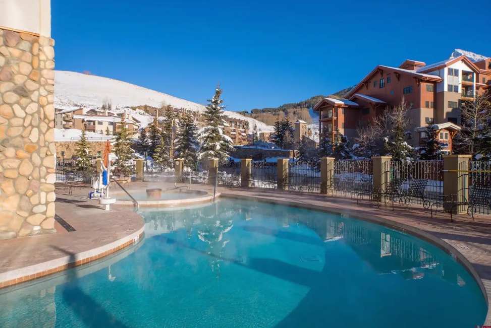 A heated outdoor pool is surrounded by a stone deck and fencing, with steam rising from the water in a winter landscape. Snow-covered trees are visible in the background, along with neighboring lodge buildings under a clear blue sky.
