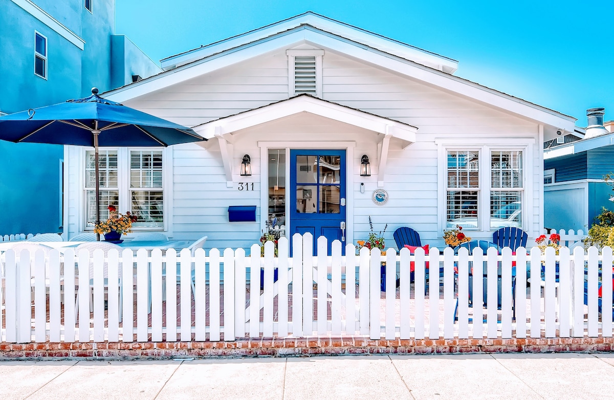 A charming cottage with bright white siding and a welcoming blue door is framed by a white picket fence. Colorful flowers adorn the front yard, while blue and white Adirondack chairs invite relaxation under a shaded umbrella.