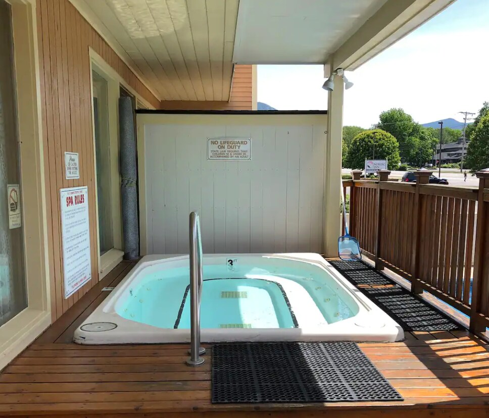 A private outdoor hot tub is situated on a wooden deck, featuring steps leading into the tub. Mats are placed around the hot tub area for safety. Scenic views of the surrounding mountains are visible in the background.