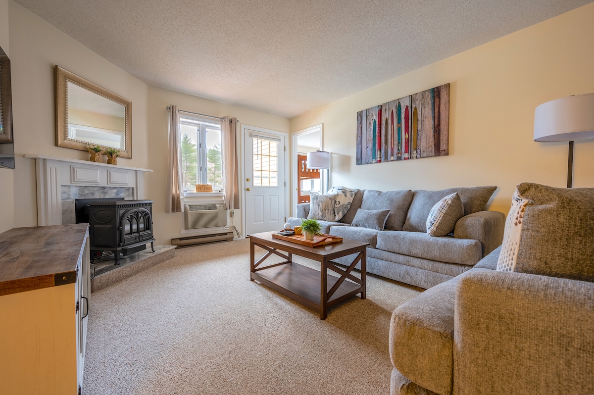 A cozy living area features a spacious gray sofa accompanied by a matching loveseat. A wooden coffee table sits centrally, adorned with a small plant. Natural light fills the room through the glass doors leading to the balcony, while a gas fireplace adds warmth to the space.