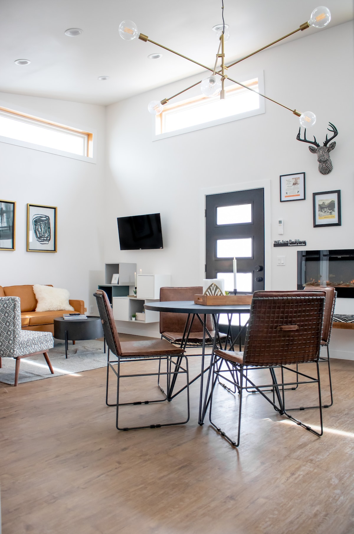 The open living space features a modern dining table surrounded by six stylish chairs. A cozy couch sits in the corner, and a TV is mounted on the wall. Accenting the area, framed artwork and a decorative wall piece add character.