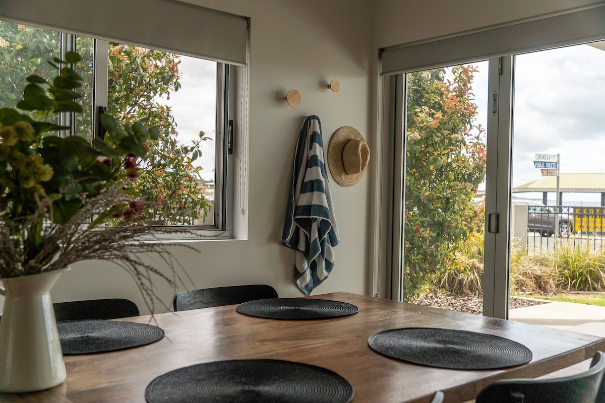 The dining area features a wooden table surrounded by black chairs, with dark placemats on the surface. Large windows allow natural light to fill the space, offering views of greenery outside. A striped towel hangs on a hook, adding a touch of casual comfort.