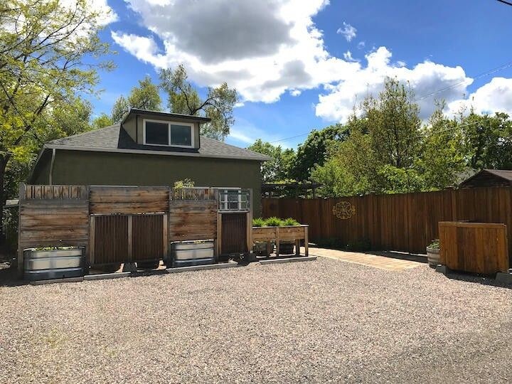 An outdoor space is visible with a gravel parking area surrounded by landscaped wooden fencing. Raised garden beds made from metal containers are placed along the fence, complemented by green foliage. A clear blue sky with scattered clouds provides natural light to the area.