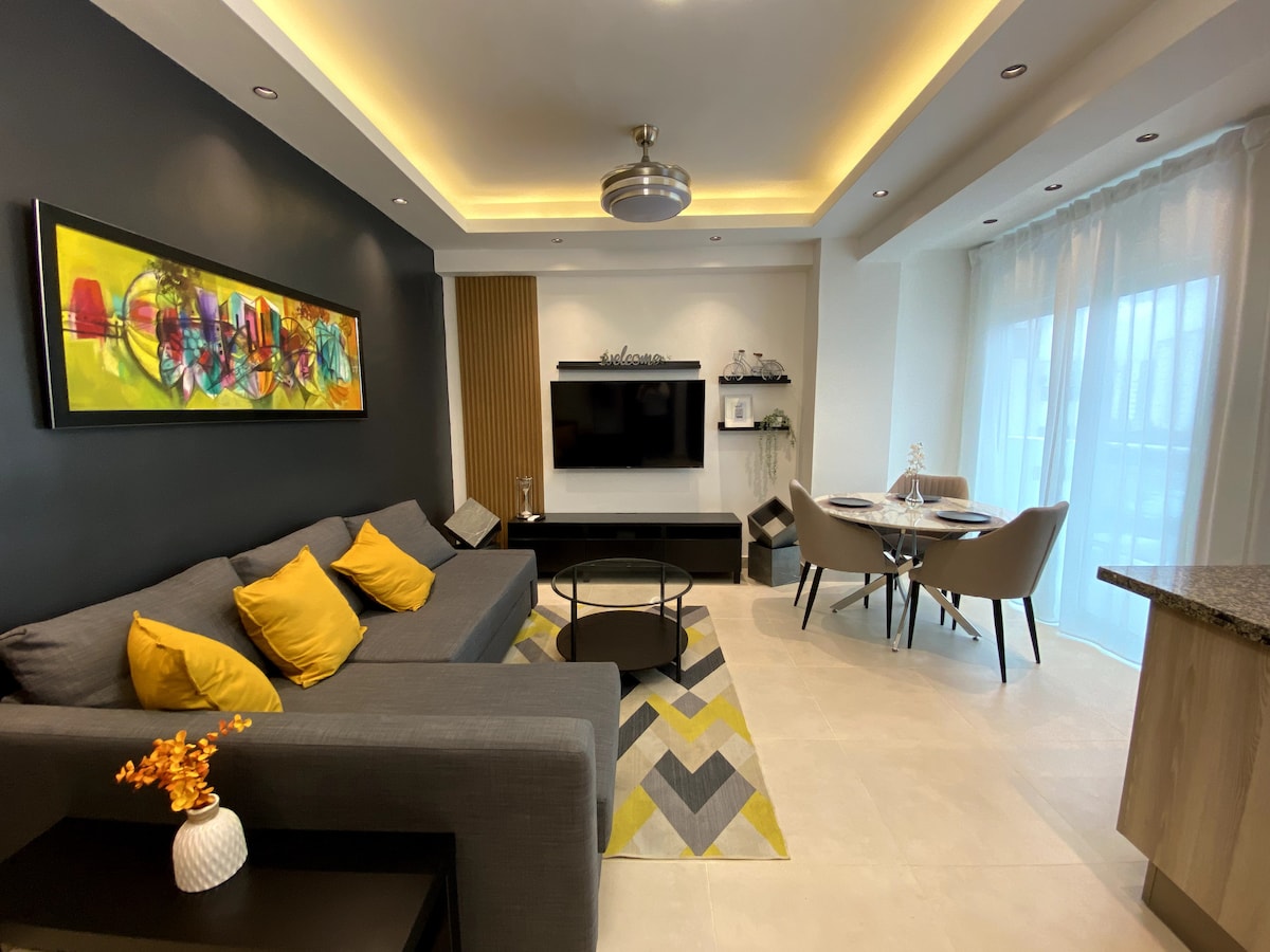 The living area features a modern gray sofa adorned with yellow accent pillows. A sleek coffee table is positioned in front of the couch, complemented by a patterned area rug. A mounted TV is visible on one wall, with a dining space featuring a round table and chairs nearby.