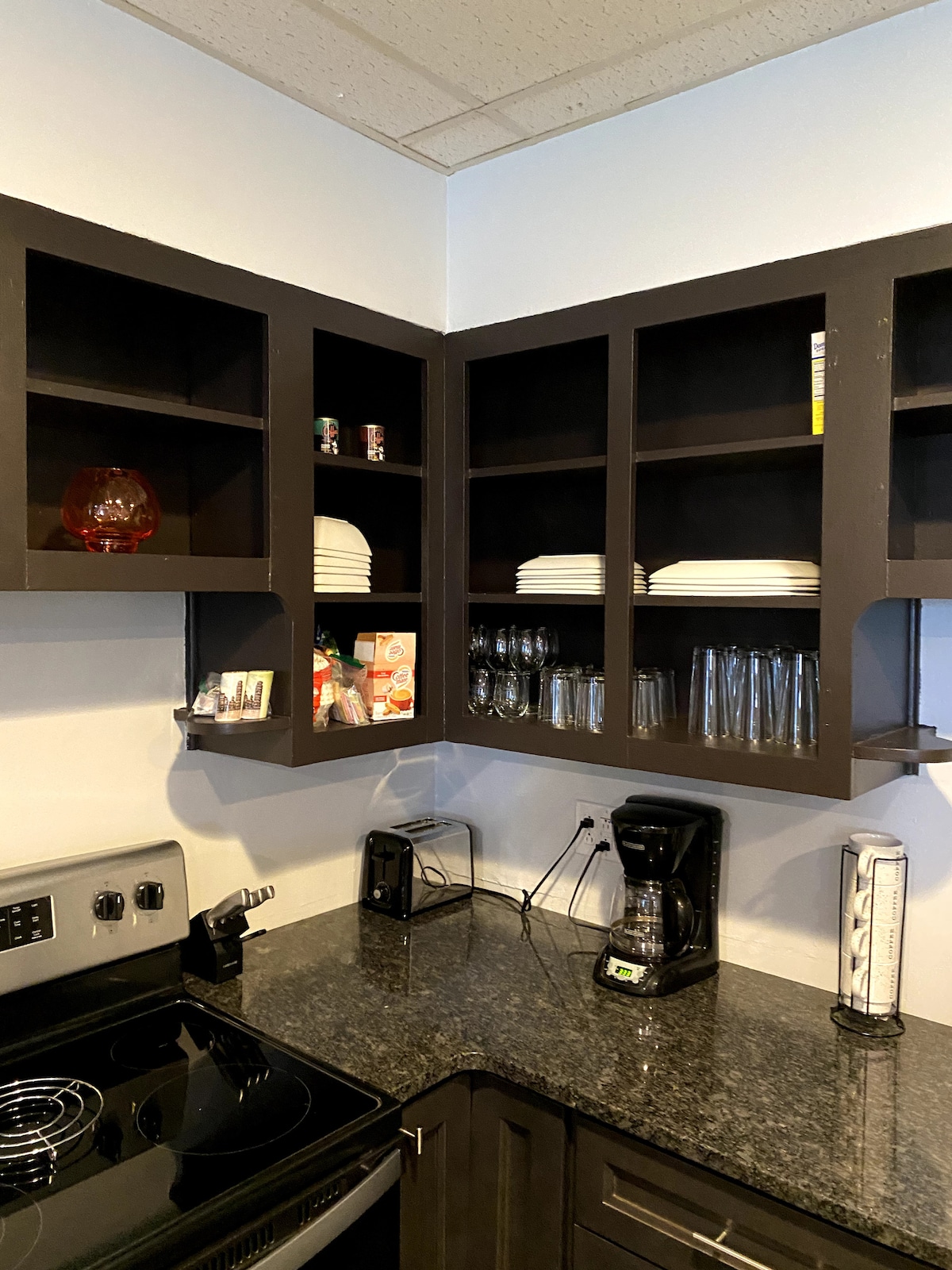 The kitchen features dark cabinetry with open shelves displaying plates and glassware. Black granite countertops complement the appliances, including a stove and coffee maker. A toaster sits on the counter, with various cooking essentials organized for convenience.