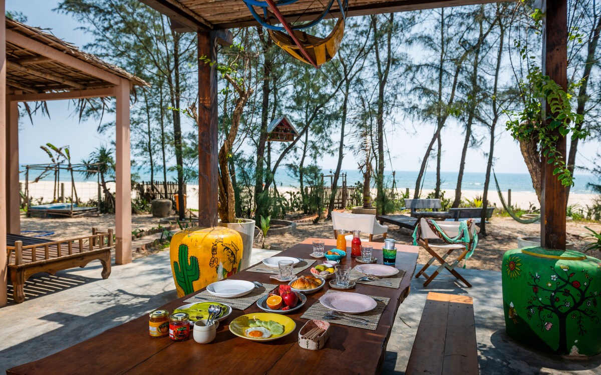 A rustic outdoor dining area features a large wooden table set with various dishes, including fruit and condiments. Colorful stools add a vibrant touch. The background showcases a beach view framed by trees, enhancing the relaxed and breezy atmosphere.