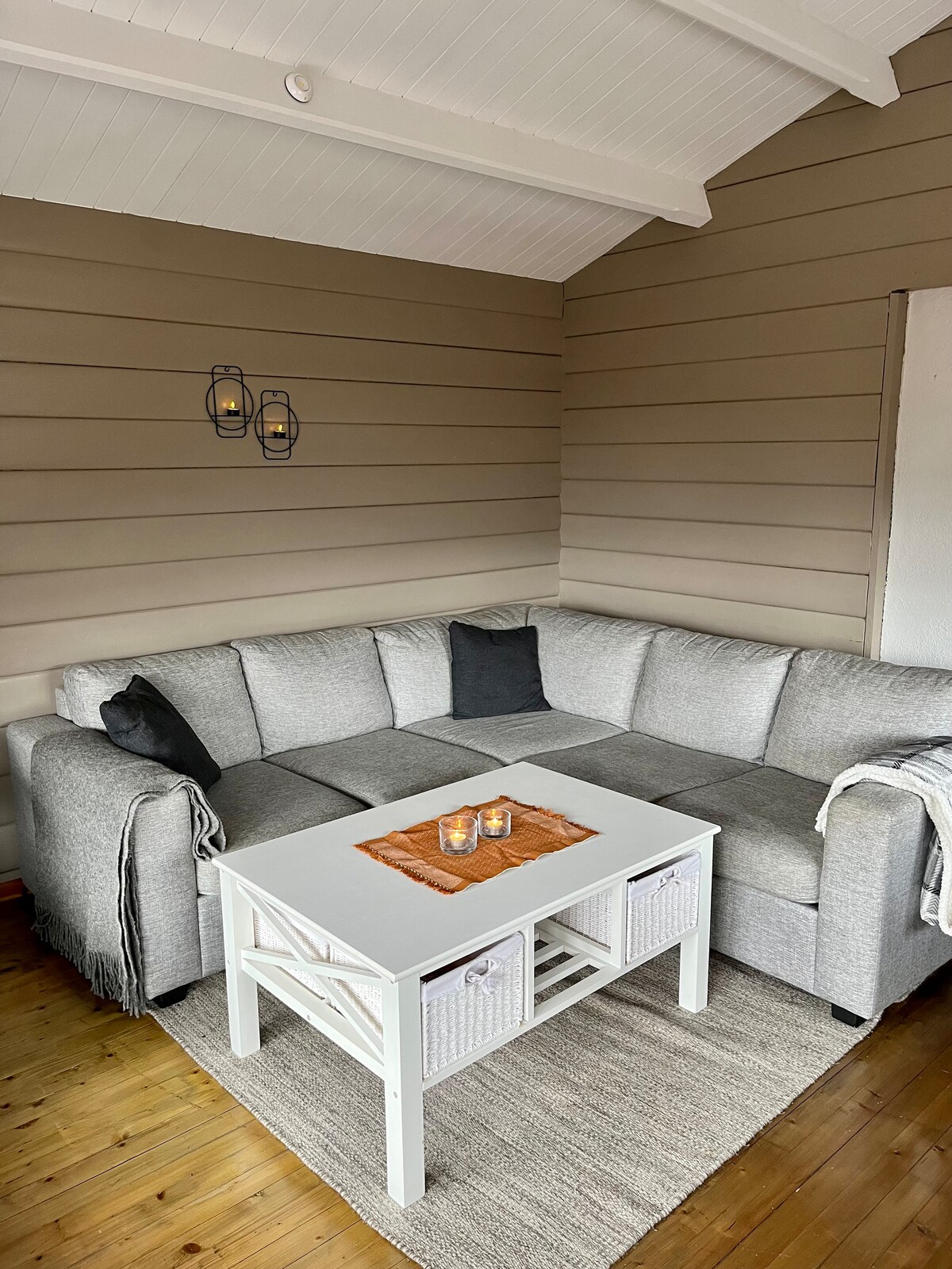 A comfortable seating area features a spacious gray and white sectional sofa adorned with dark cushions. A white coffee table, accented with woven baskets, sits in the center, accompanied by candles on a decorative tray. Warm wood flooring enhances the inviting atmosphere.