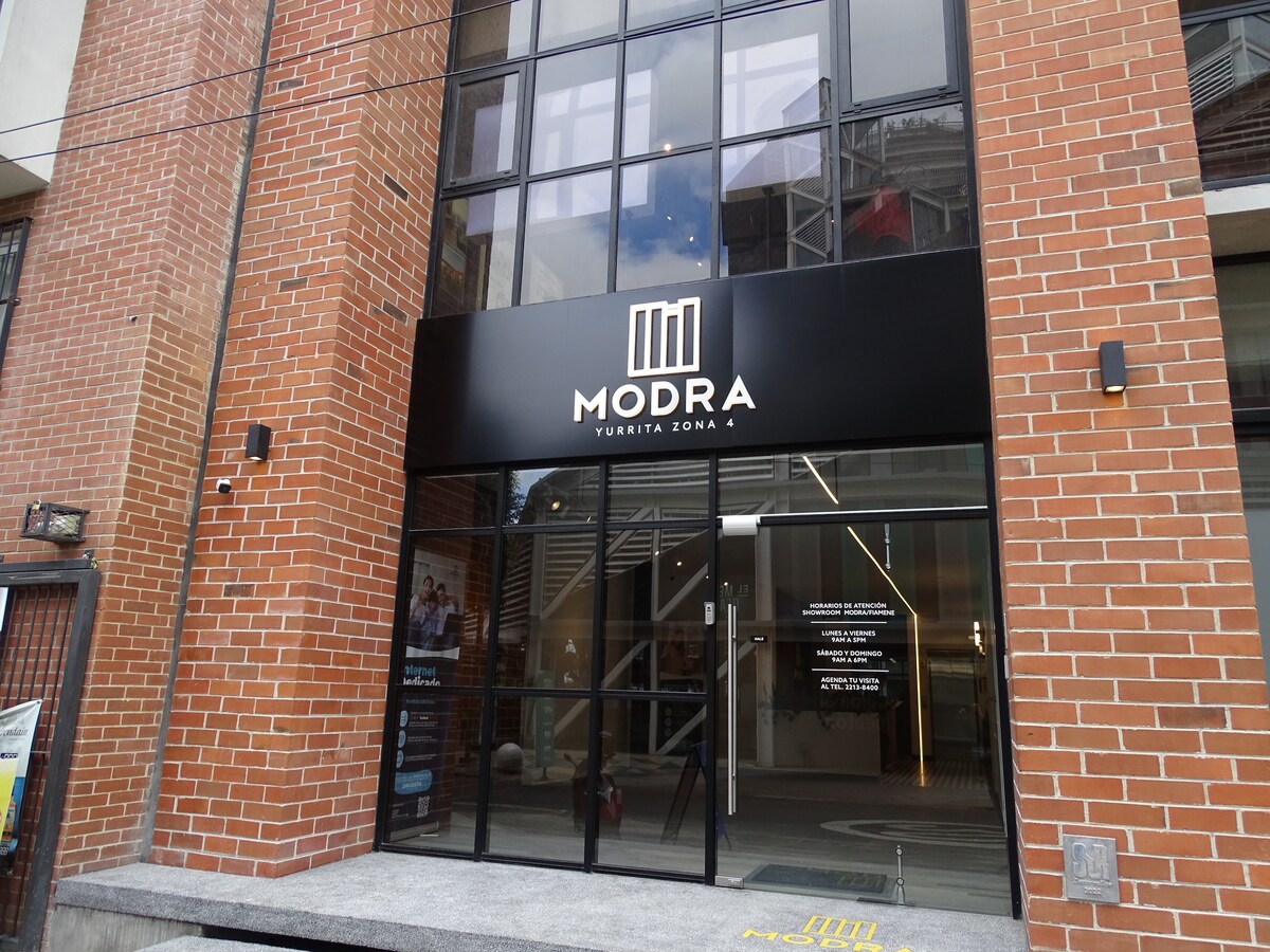 The entrance of the MODRA building is displayed, featuring a modern design with large glass doors framed by brick walls. The black sign prominently features the name 'MODRA,' while the overall façade suggests a contemporary architectural style.