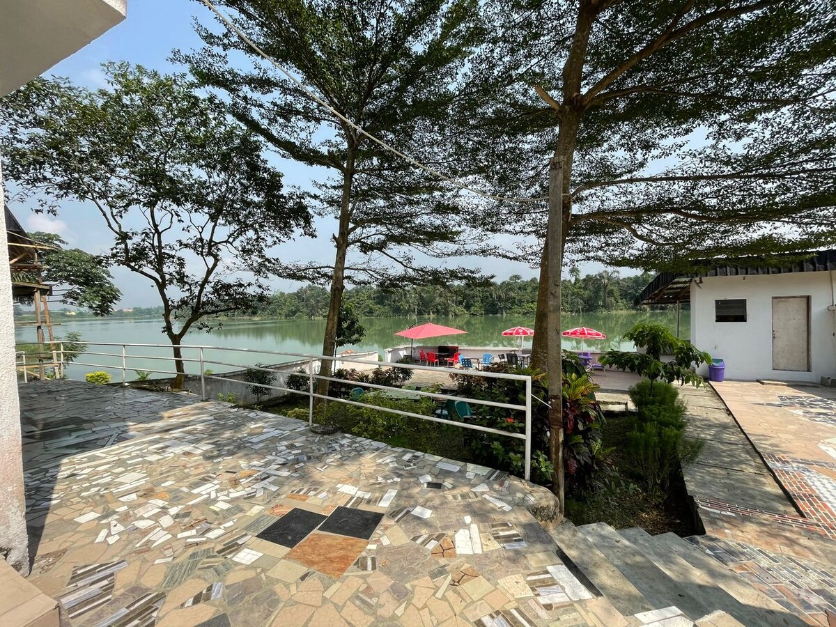 A serene view of the riverfront, framed by tall trees. A paved area with varied stone patterns leads to the water. Seated areas with umbrellas are visible near the water’s edge, creating a tranquil outdoor space for relaxation.