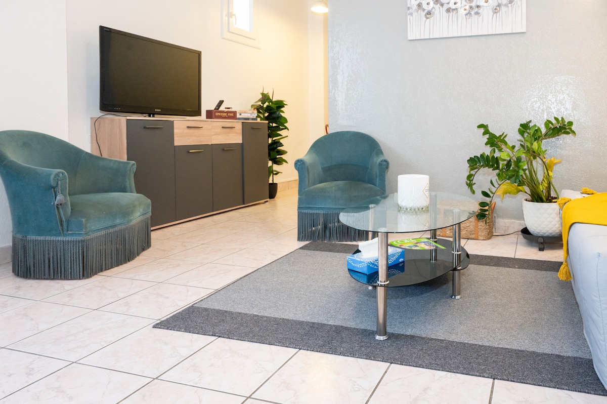A welcoming living area is presented with two soft armchairs in teal and a stylish glass coffee table at its center. A large television is positioned against the wall, complemented by a sleek cabinet. Potted plants add a touch of greenery to the space.