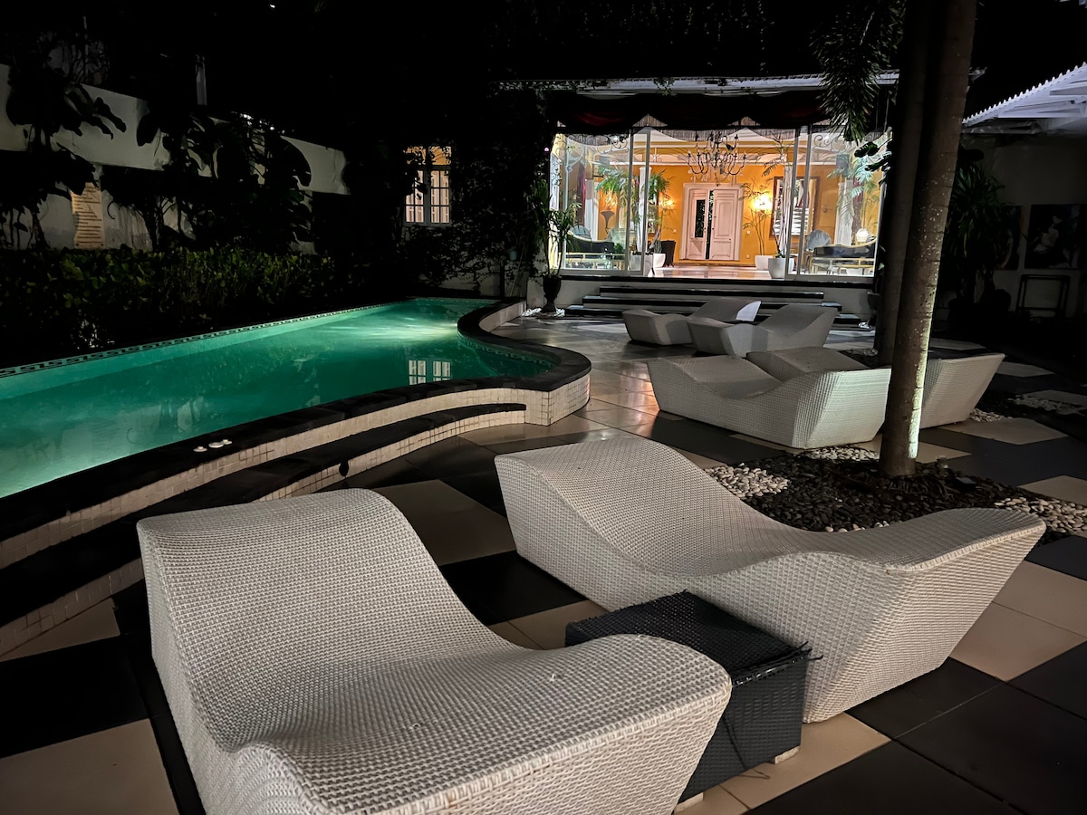 A beautifully illuminated pool area features a curvy swimming pool surrounded by lounge chairs. Lush greenery adds a serene touch, while the glass doors of the main living space are visible in the background, revealing a well-decorated interior.