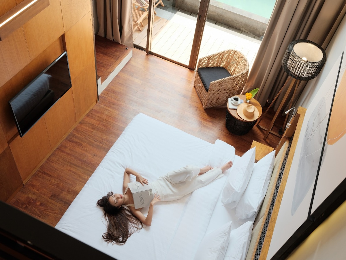 A spacious bedroom features a large bed with neatly arranged pillows. Natural light floods the room through large glass doors, which open to a private area. A cozy wicker chair and a small table enhance the space, creating a relaxing environment.
