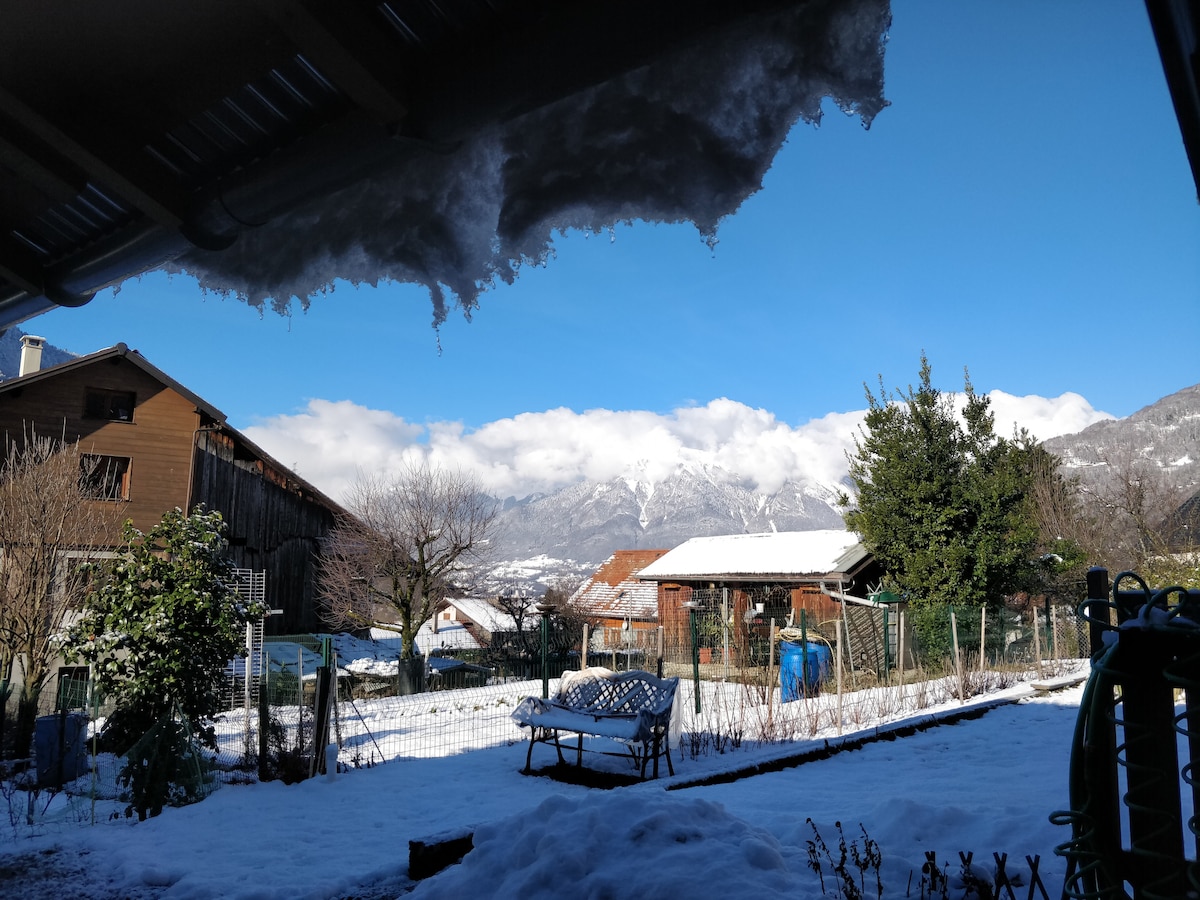 A winter landscape is visible from a covered outdoor area, featuring snow-covered ground and a view of distant mountains shrouded in clouds. Nearby, residential buildings and trees are framed against a clear blue sky, enhancing the serene atmosphere of the scene.