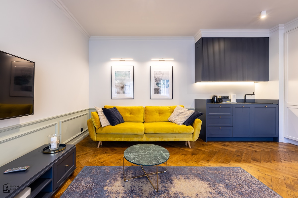 A living area is presented with a vibrant yellow sofa, complemented by a dark blue kitchenette. Two framed artworks hang above the seating area. A round coffee table with a marble surface occupies the center of the room, positioned on a textured rug.
