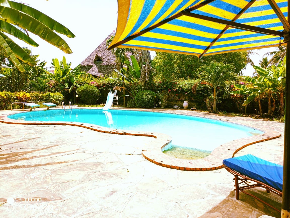 A swimming pool is surrounded by a stone patio and vibrant tropical plants. A blue sunbed is positioned nearby, and a shaded area is created by a large yellow and blue striped umbrella. A waterslide is visible at one end of the pool.