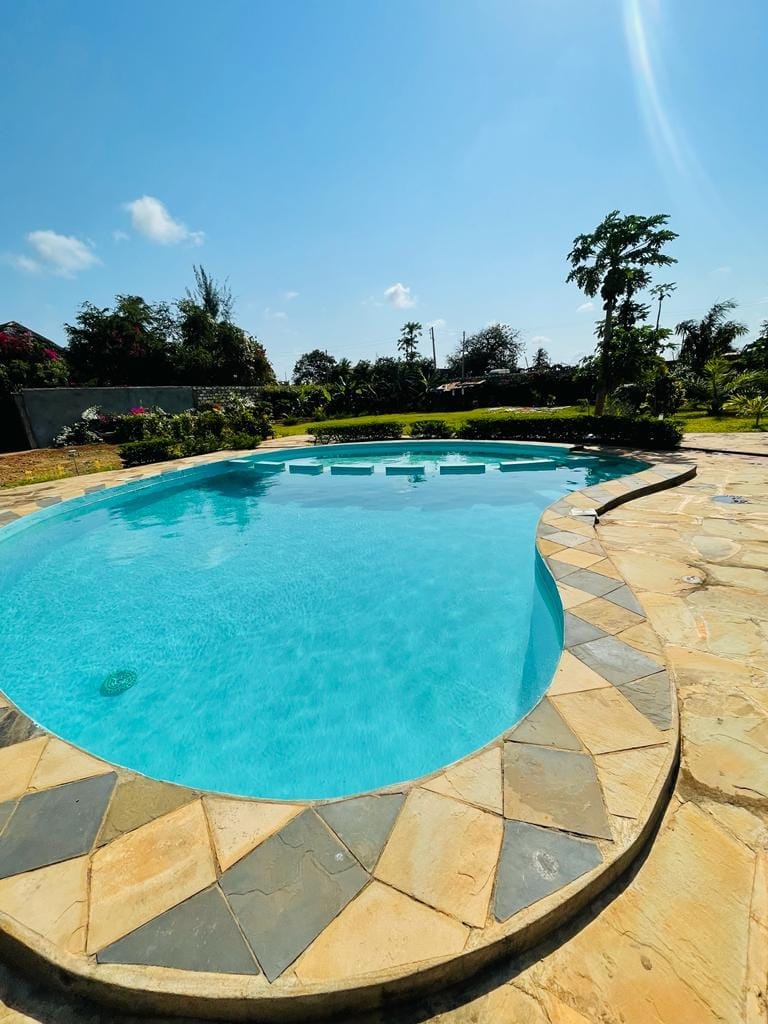 A clear swimming pool is surrounded by a natural stone deck, featuring a curved edge that blends into a spacious garden area. Lush greenery and blooming flowers are visible in the background, under a bright blue sky.