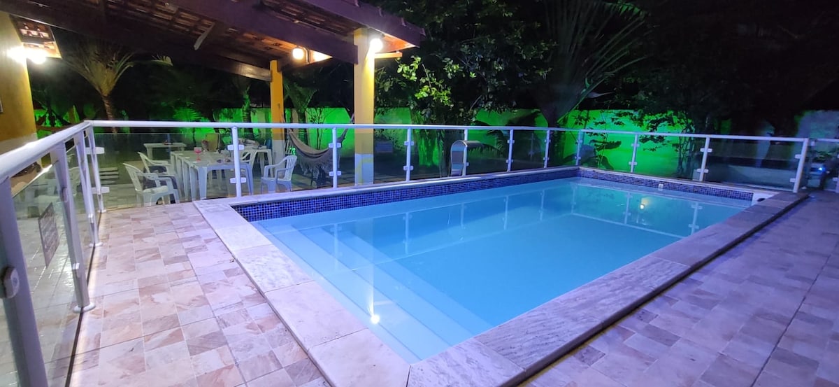 A well-lit pool area is surrounded by a safety glass railing, with a stone patio featuring lounge chairs and a dining set. Lush greenery provides a serene backdrop to the tranquil water, creating an inviting space for relaxation.