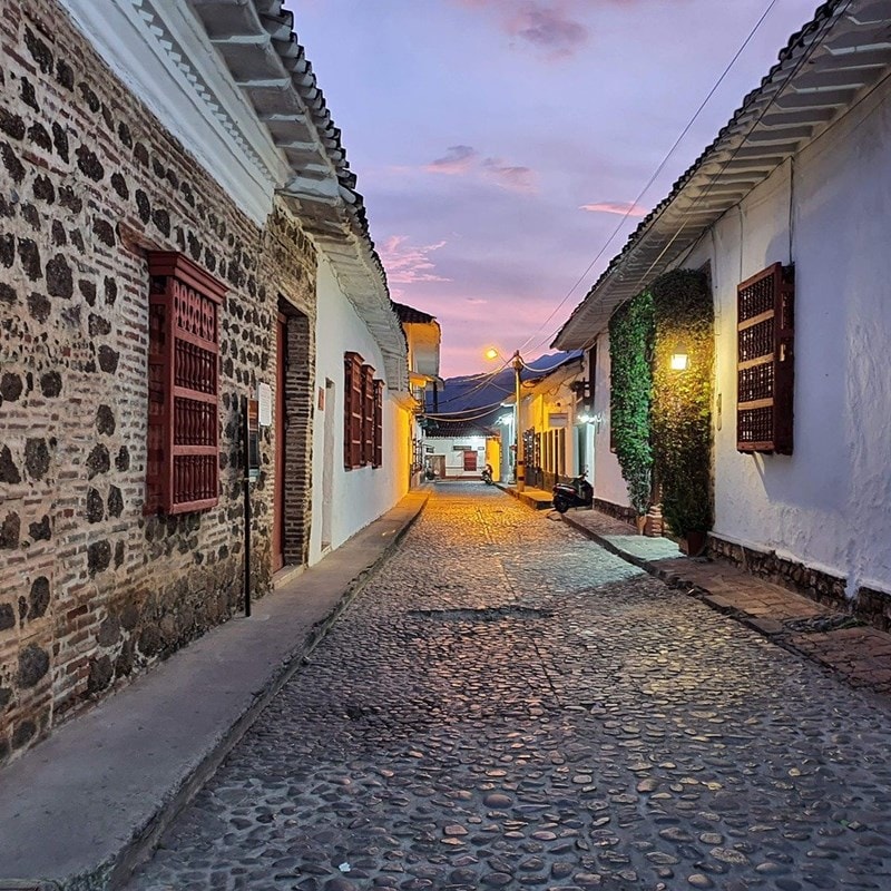 A cobblestone street is bordered by charming colonial buildings with whitewashed walls and red wooden windows. The soft glow of street lamps highlights the evening sky, where colors transition from orange to purple as dusk settles, creating a peaceful ambiance.