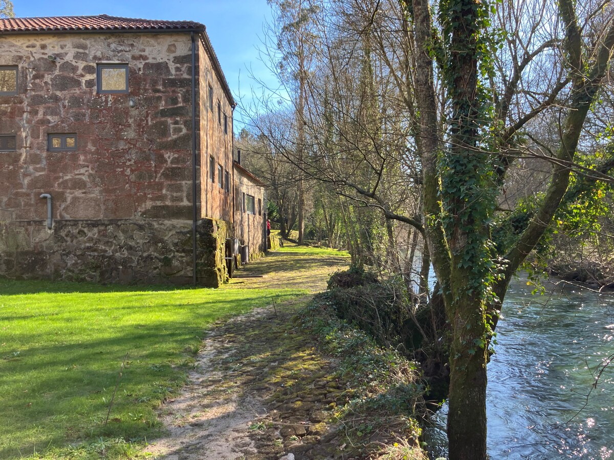 A serene pathway runs alongside a flowing river, framed by lush greenery. An old stone building with rustic charm stands nearby, showcasing large windows that reflect the surrounding nature. The riverbank is gently sloped, inviting peaceful contemplation by the waterside.