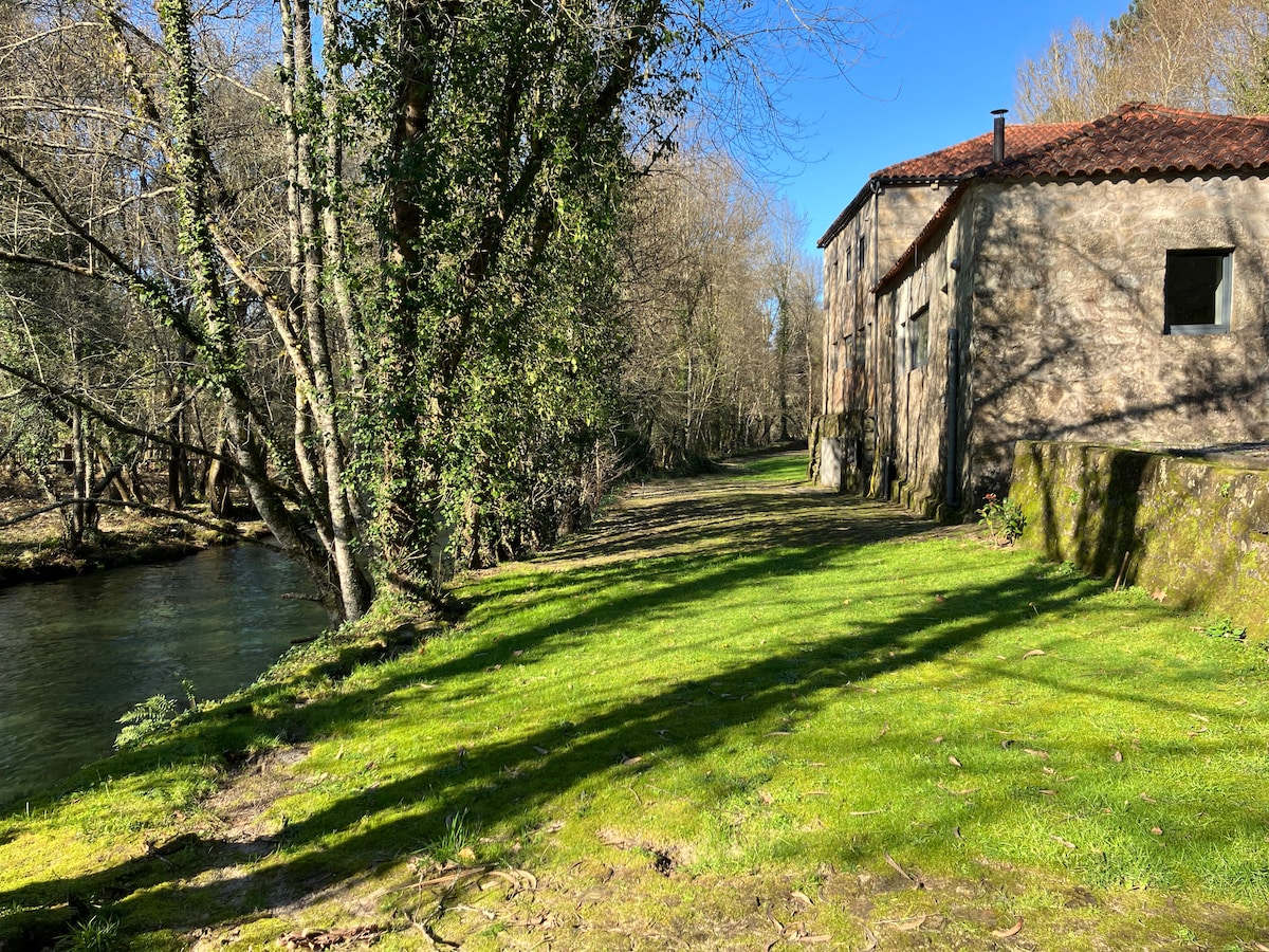 A serene green area extends alongside the River Vez, bordered by trees casting gentle shadows. A rustic stone building is seen in the background, with a roof covered in terracotta tiles and a window that allows glimpses of the surrounding natural landscape.